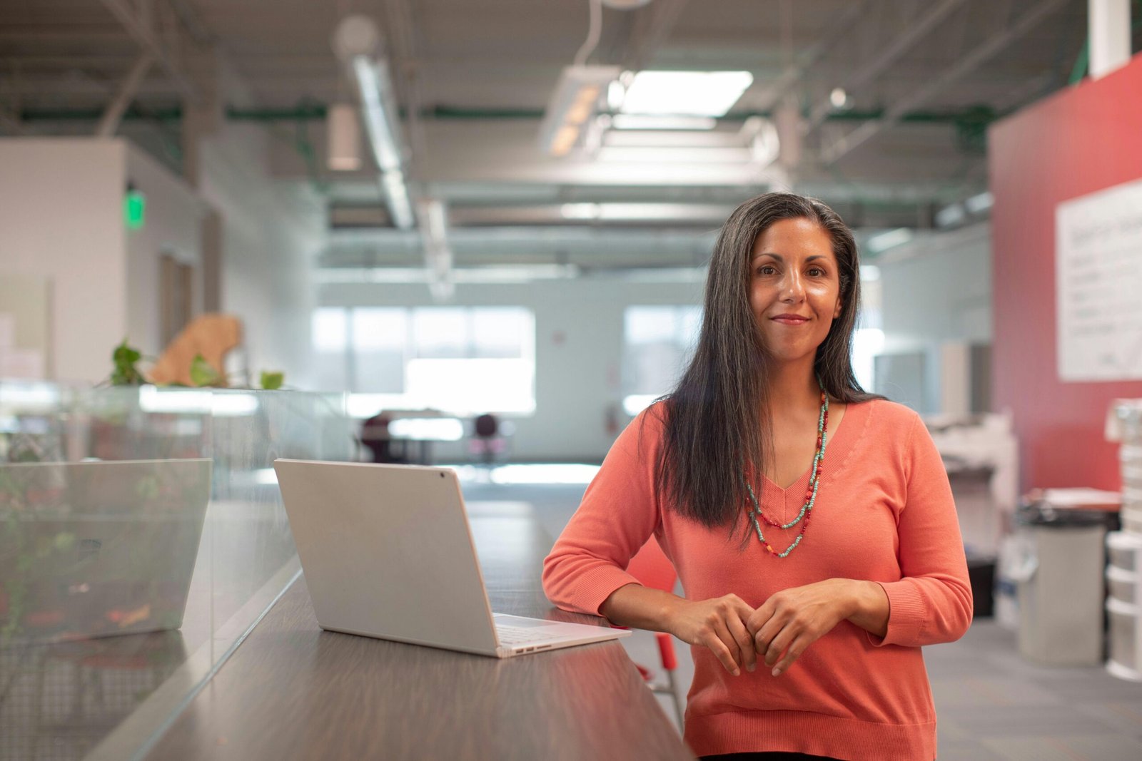 Smiling woman in a bright office, standing by her laptop, exuding confidence.